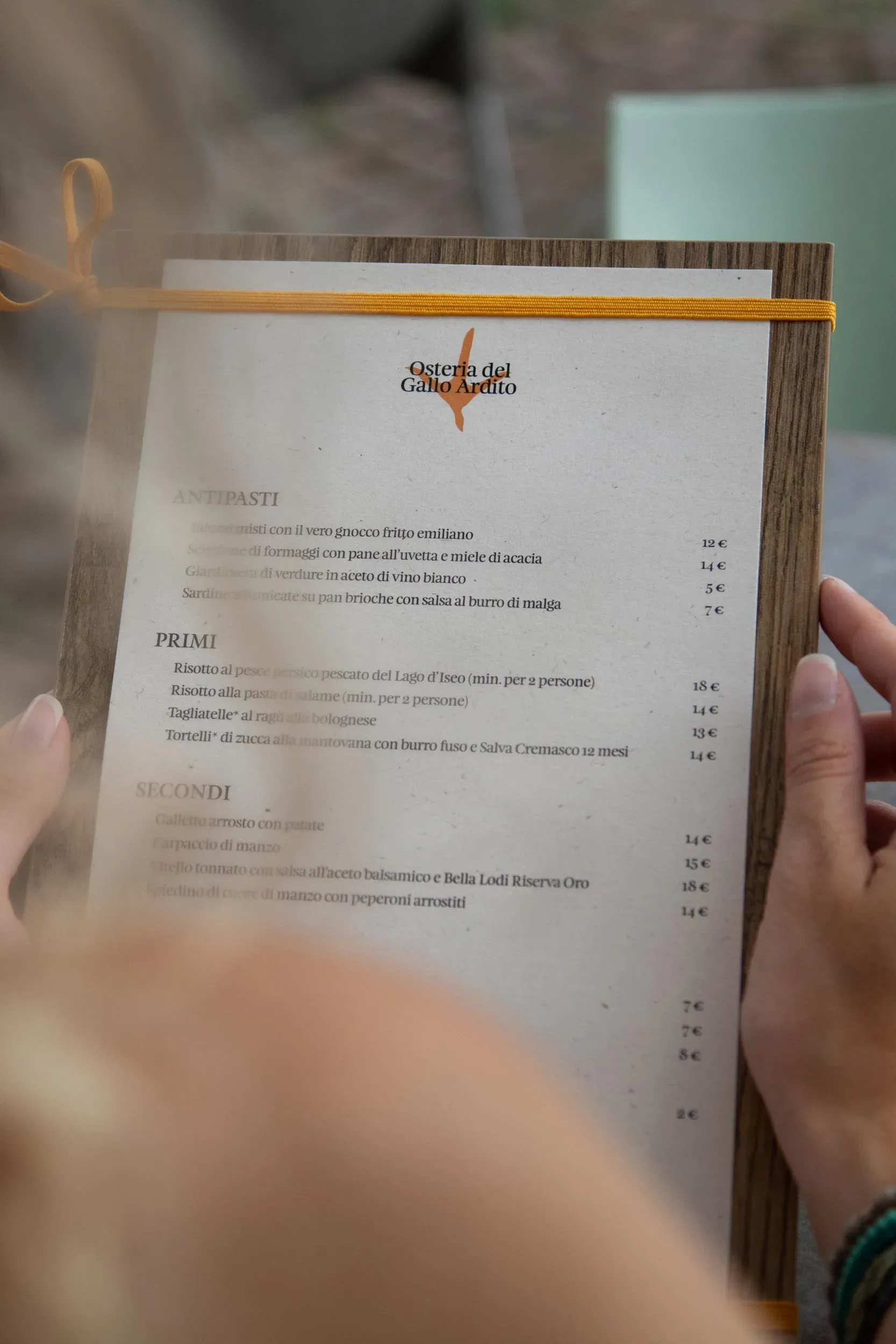 Close-up of the Osteria del Gallo Ardito menu held by a person, showing traditional Italian dishes printed on textured paper with a wooden frame and orange cord | Carlotta Bacchini