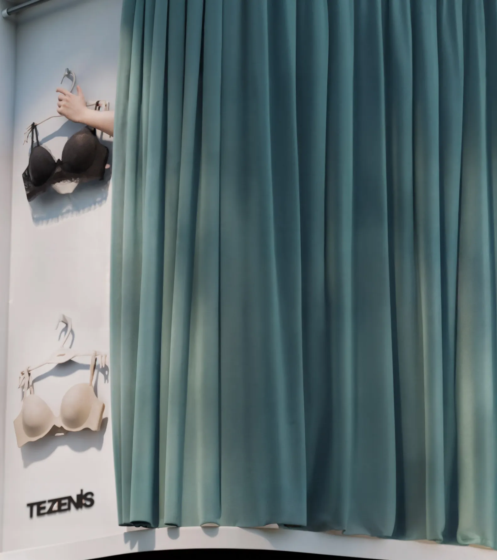 Tezenis billboard detail with a hand selecting a black bra from a display, next to a teal curtain partially revealing a white bra on the wall. | Carlotta Bacchini