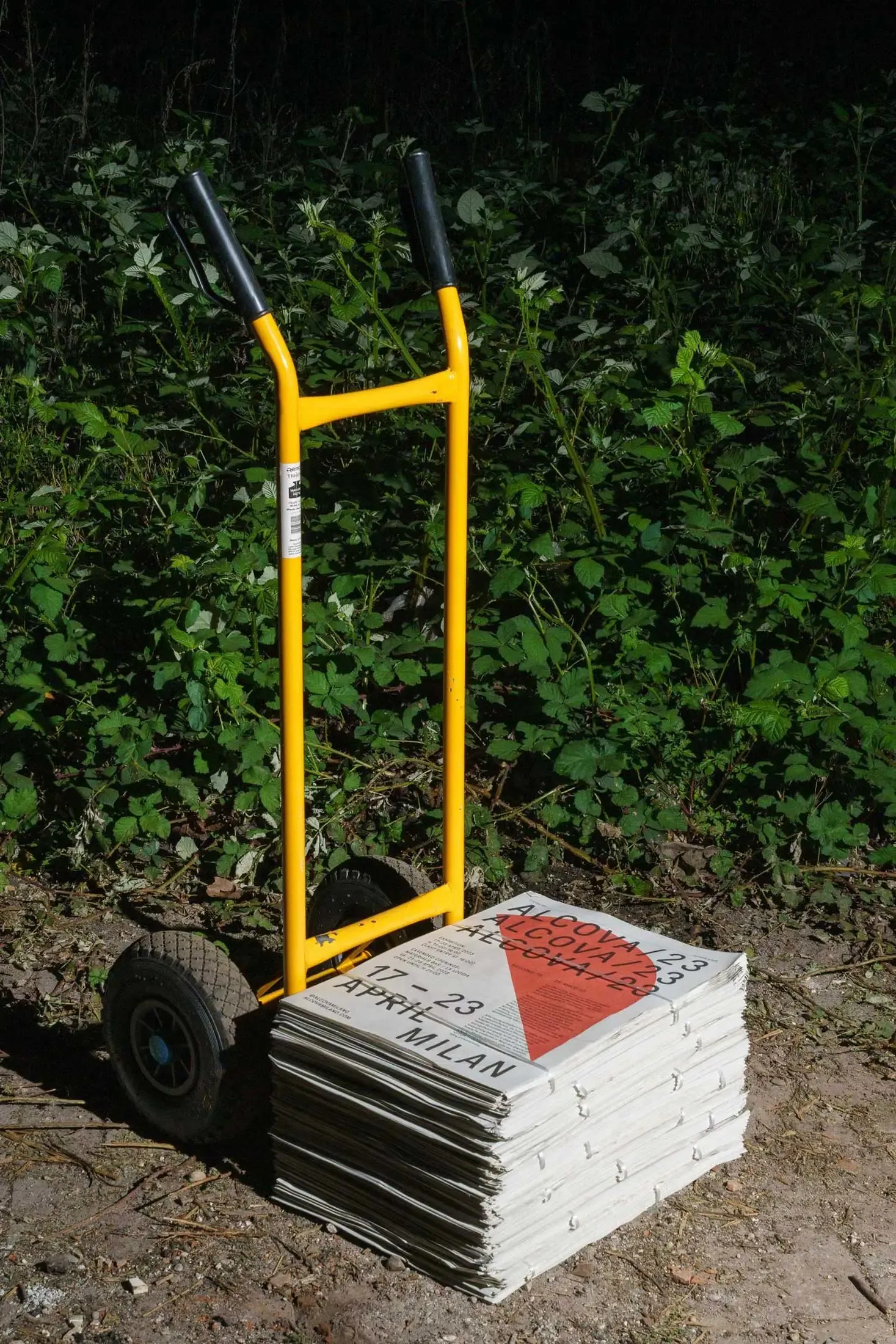 Stack of Alcova 23 fold-out newspaper maps with black and orange geometric design placed on a yellow hand truck, photographed outdoors | Carlotta Bacchini
