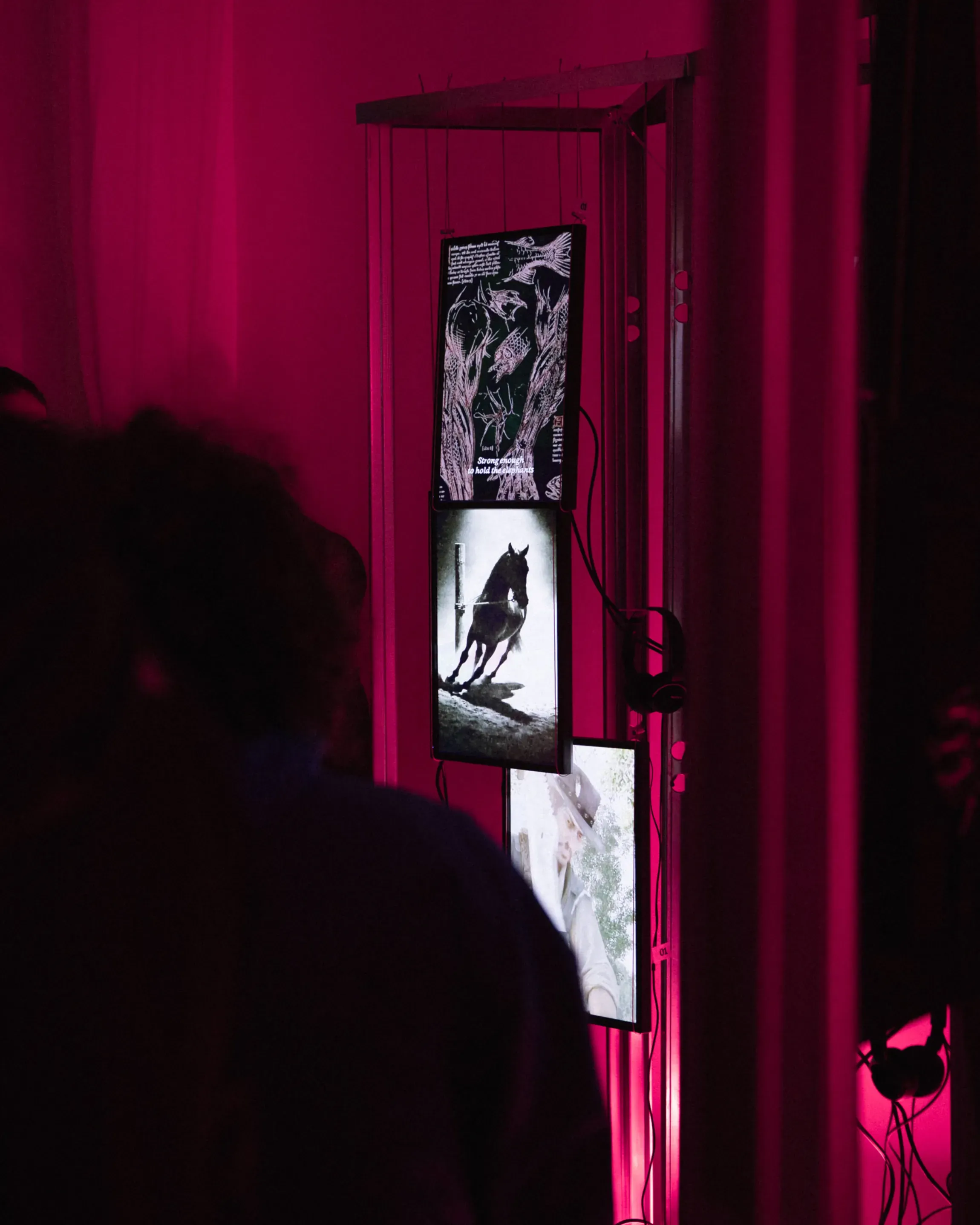 Detail of suspended monitors and headphones in An Ecology of Beasts exhibition, showing collective video works inspired by medieval bestiaries, pink lit installation