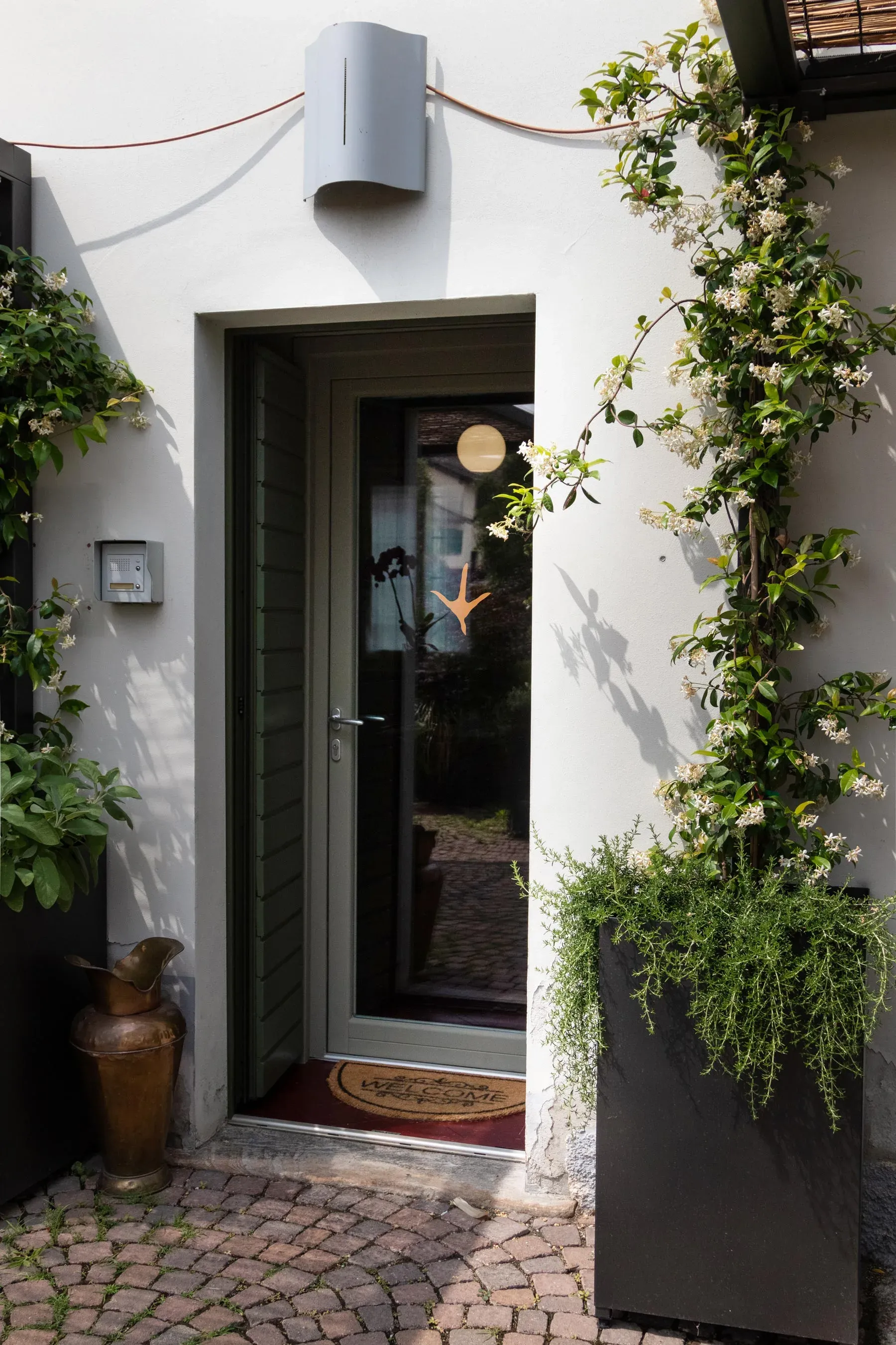 Entrance of Osteria del Gallo Ardito with logo on glass door, surrounded by flowering plants and traditional stone pavement | Carlotta Bacchini