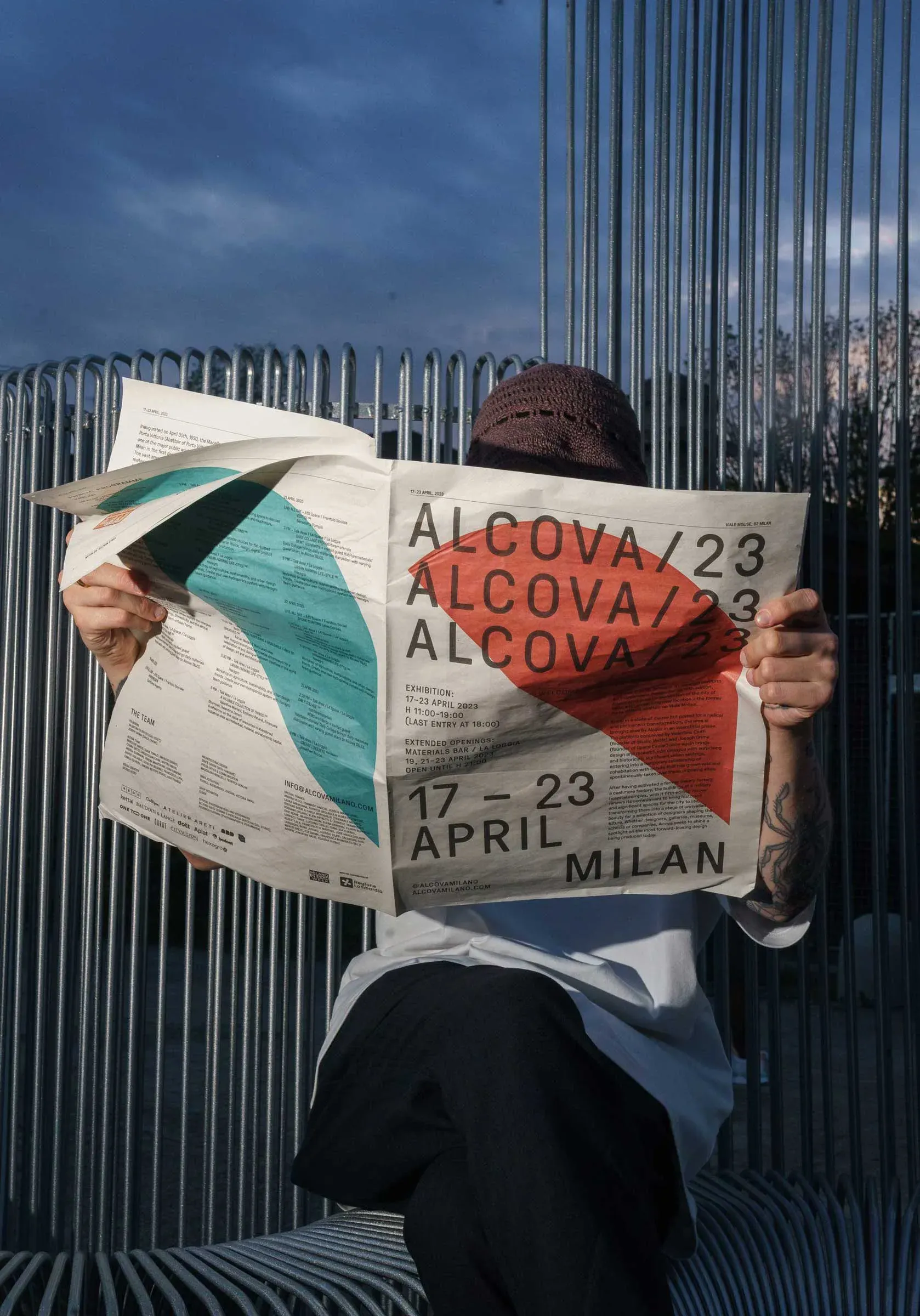Person reading the Alcova 23 newspaper with bold black and orange geometric design, folded open to show event details, photographed outdoors. | Carlotta Bacchini