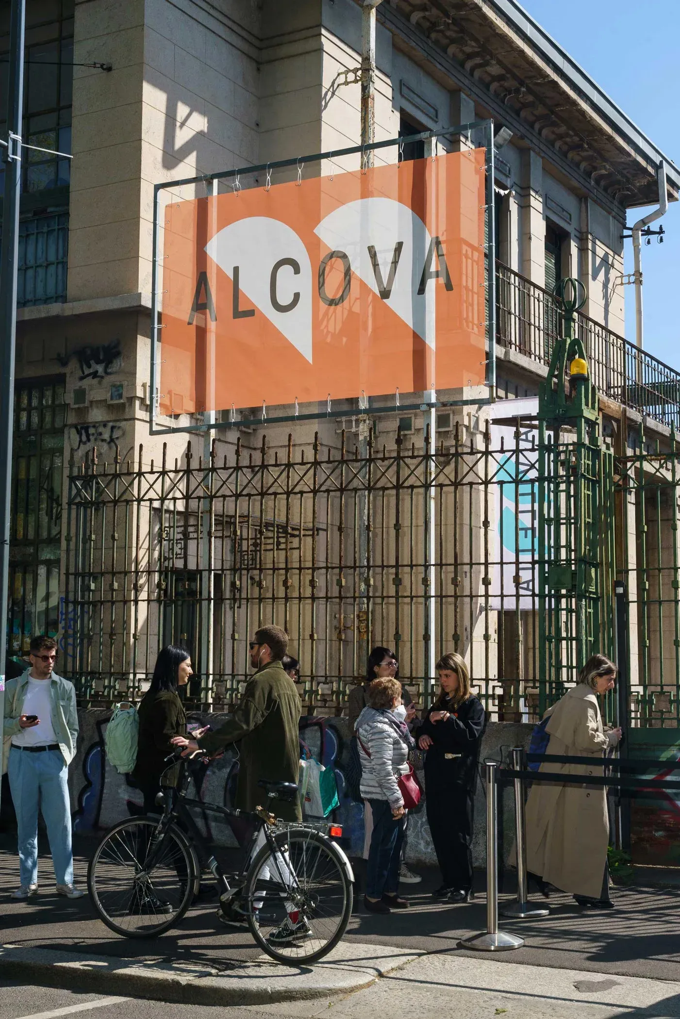 Outdoor photo of the Alcova exhibition banner in orange and white colors, hanging on the former Porta Vittoria abbattoir facade