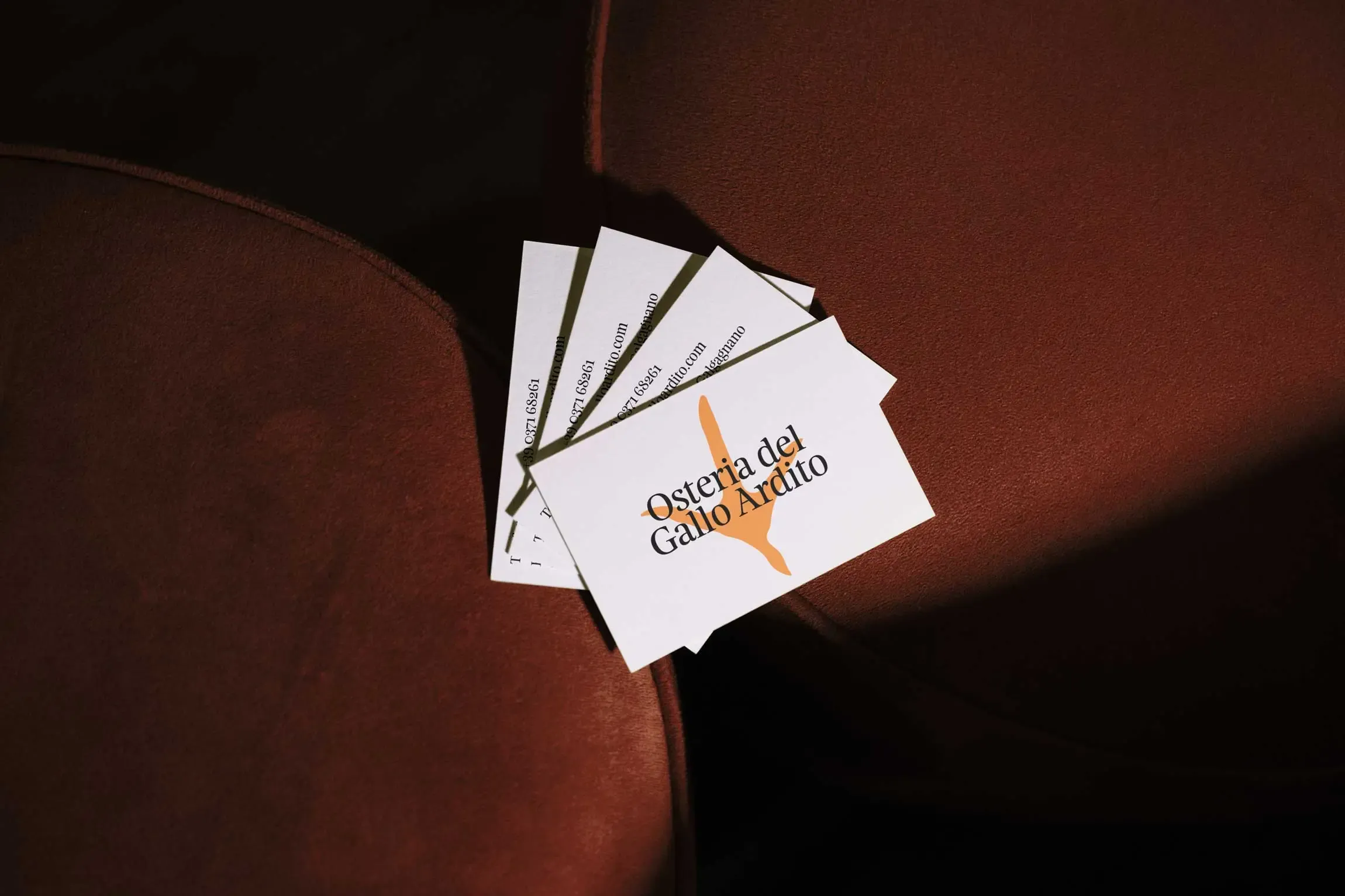 Set of business cards for Osteria del Gallo Ardito displayed on a red velvet surface, featuring the restaurant’s name and orange rooster foot logo | Carlotta Bacchini