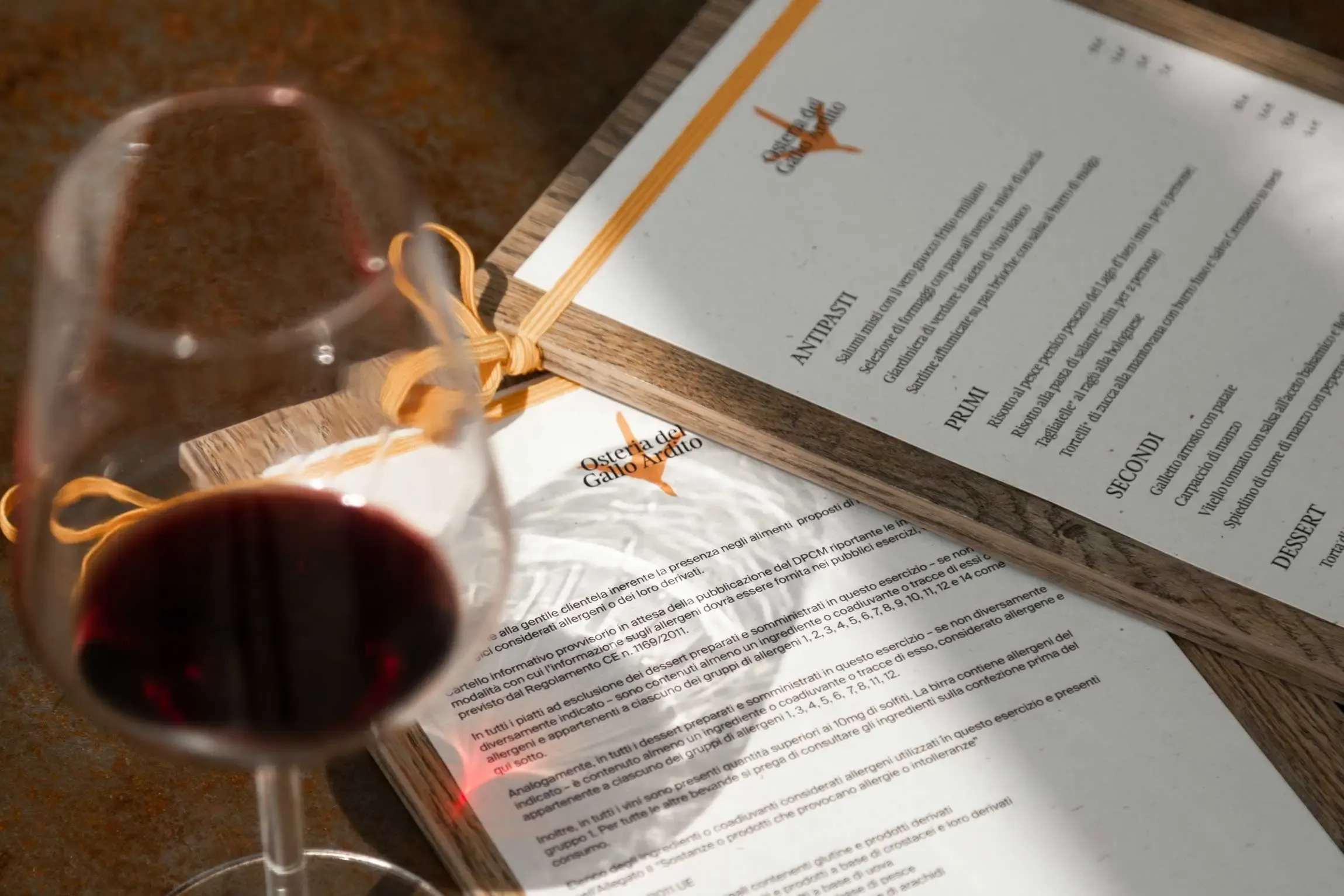 Close-up of Osteria del Gallo Ardito’s printed menu framed in wood with an orange cord, next to a glass of red wine casting a shadow on the paper| Carlotta Bacchini